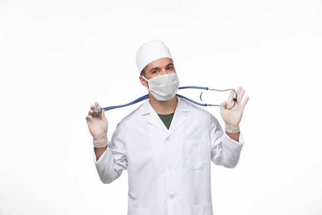 专业正面图：男医生穿着防毒服 戴着口罩 手持听诊器 在白墙病毒病大流行听诊器套装视图