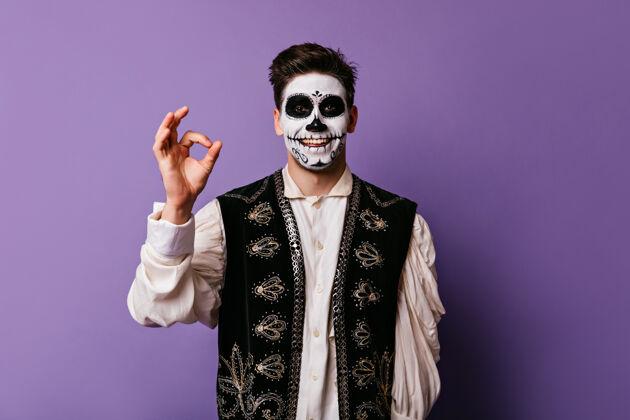 文化快乐的僵尸男人在紫色的墙上微笑快乐的年轻人带着可怕的化妆品在万圣节摆出了一副好的姿势骷髅传统室内