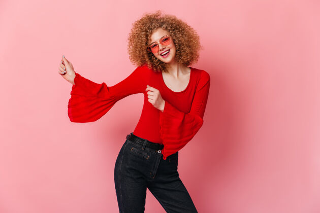 红色衬衫在粉色空间里跳舞的卷曲可爱的女人戴着粉色眼镜 穿着红色衬衫和牛仔裤的金发女郎的肖像衣服优雅表情