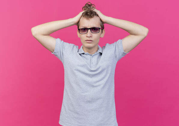 眼镜一个戴着黑眼镜 穿着灰色马球衫的失望的年轻人 站在粉红色的墙上摸着他的头 感到困惑头摸着站着