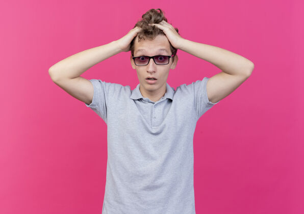 失望一个戴着黑眼镜 穿着灰色马球衫的失望的年轻人 站在粉红色的墙上摸着他的头 感到困惑男人马球年轻