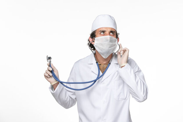 使用正面图：男医生穿着医疗服 戴着口罩以防感染冠状病毒-使用听诊器对白墙病毒喷溅冠状病毒大流行面罩防护喷溅