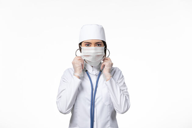 面罩正面图女医生穿着白色无菌医疗服 戴口罩因冠状病毒-使用听诊器对白墙病病毒冠状病毒-大流行性疾病使用消毒冠状病毒