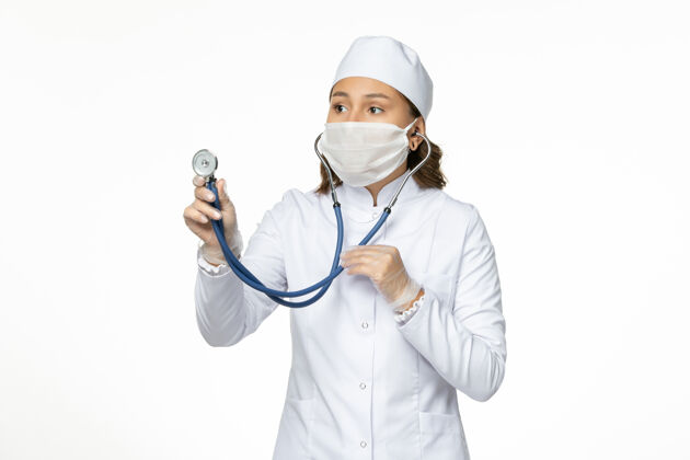 检查前视图穿着白色医疗服和面罩的女医生在白色办公桌上用听诊器检查心脏状况潘多米病毒病药物女性医学状况