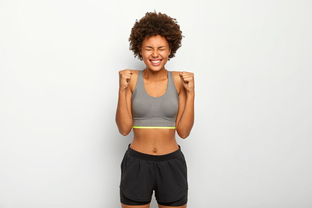 健康精力充沛的运动女性为胜利而欢欣鼓舞 举起紧握的拳头 笑容灿烂 穿着运动内衣 笑容灿烂 与白色背景隔绝 锻炼运动文胸