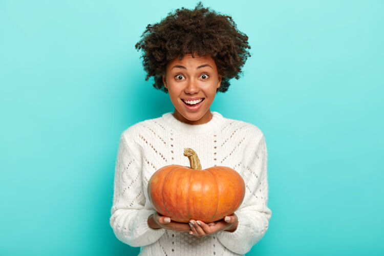 牙关快乐的非洲裔美国女人抱着橙色的南瓜 穿着白色的毛衣 温柔地微笑 孤立在蓝色的墙上年轻秋天欢乐