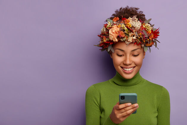 自由空间秋高气爽的模特戴着装饰性的秋日花环 专注于智能手机设备 在线阅读好消息 有着幸福的表情 模特们翻过紫色的工作室墙壁拿着手机信息