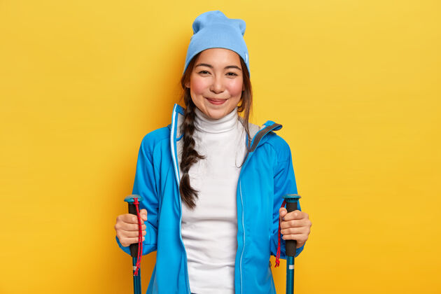 私人教练积极的年轻黑发女子喜欢北欧散步 手持登山杖 在森林小道上训练 戴蓝色帽子 穿夹克和白色高领毛衣 在黄色背景下摆姿势徒步旅行和露营运动蓝色举行