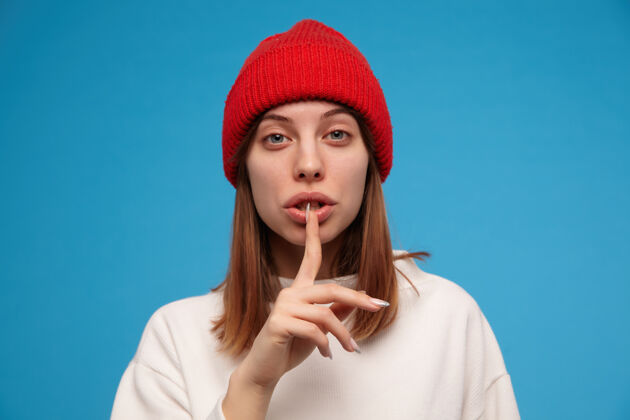 嘴唇十几岁的女孩 深色头发的女人穿白色毛衣 戴红色帽子显示沉默标志 保持安静人的概念特写镜头 隔离在蓝色的墙上年轻黑发室内