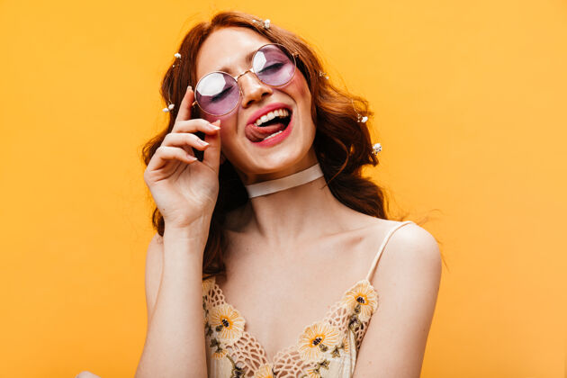 漂亮戴着淡紫色太阳镜的淘气女人舔着嘴唇 摆出橙色背景的姿势微笑外观模特