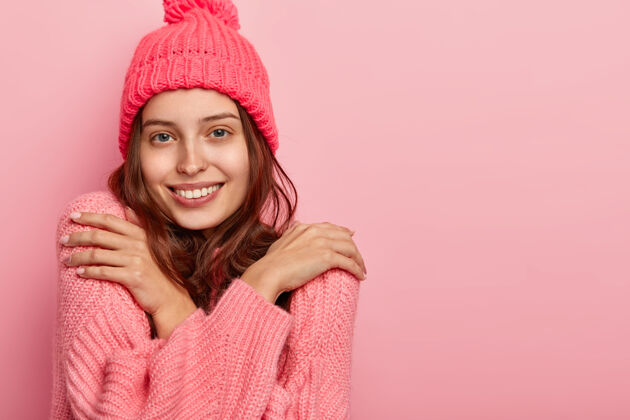 爱一个满意的微笑的女人穿着冬季针织毛衣温暖的照片 双臂交叉在胸前 抚摸着肩膀 有吸引力的样子 在粉色背景下摆姿势 旁边有自由的空间粉色休闲服装