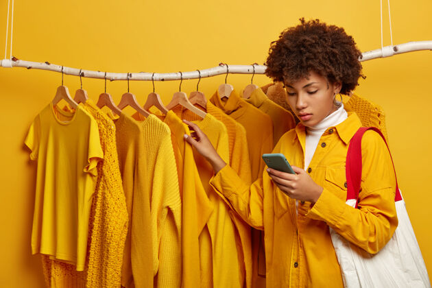 室内严肃的卷发女买家拎着包 拿起黄色的衣服 专注于智能手机 在时尚服装架旁摆姿势 购买黄色年轻设备