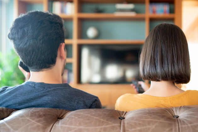 冠状病毒年轻夫妇坐在家里的沙发上一起看电视剧或电影的照片新的正常生活方式概念观看年轻在一起