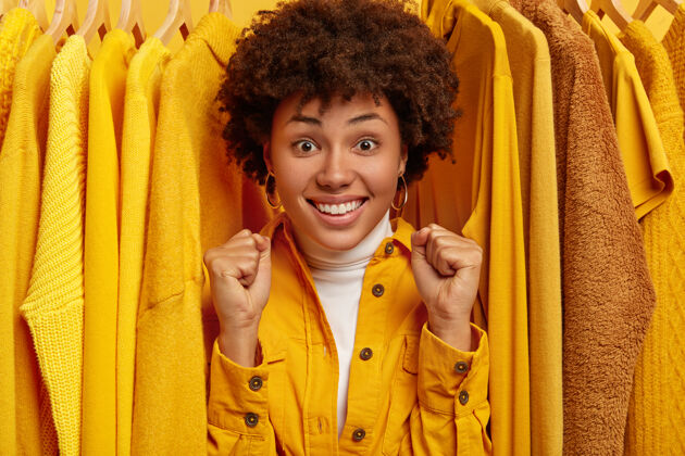 满意喜出望外的黑皮肤女人握紧拳头 欢庆衣柜的更新换代 站在衣架上的黄色服装之间 站在试衣间人与时尚的概念一个黄色购物狂