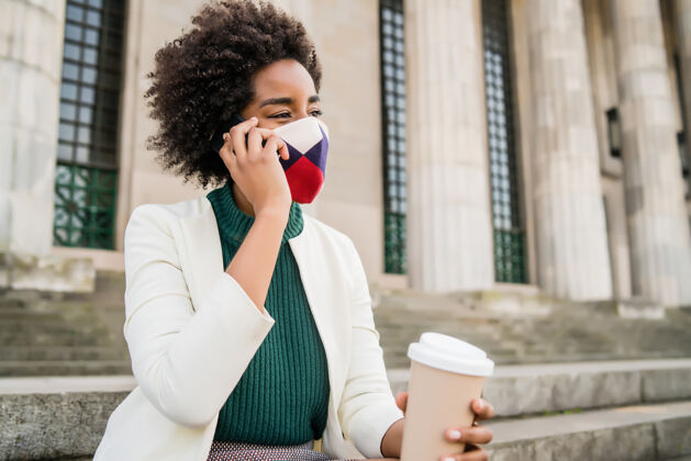 防护面罩一位非洲裔女商人戴着防护面具 坐在街上的楼梯上打电话商业和城市概念人手持应用程序