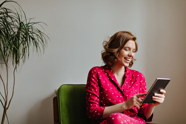 黑发坐在扶手椅上使用数码平板电脑的笑脸女人穿着睡衣的漂亮女人拿着小玩意微笑的室内照片房间棕色头发欢呼