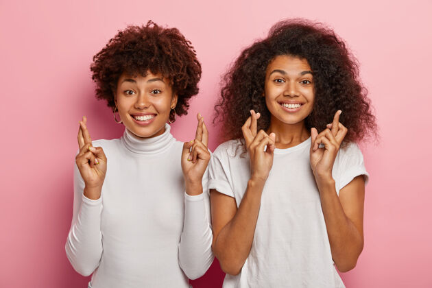 女人两个快乐的卷发女人手牵手祈求好运 穿着白色的衣服 开心地微笑 期待考试成绩 站在粉红色的背景下朋友希望标志
