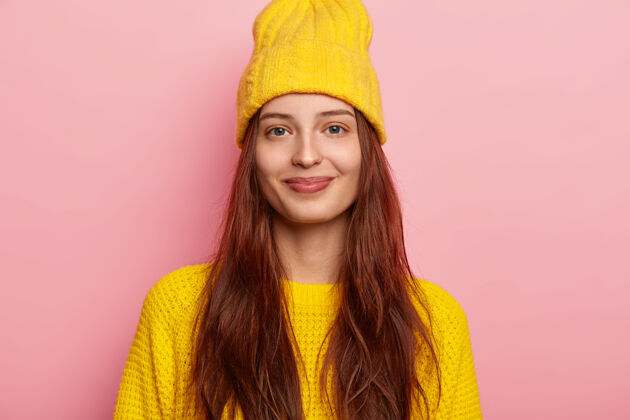 一件满意可爱的年轻女模特 戴着时髦的黄色帽子 穿着针织毛衣 留着长长的头发 在粉色背景下摆出姿势 展现出她冬日的装扮 带着温柔的微笑直视镜头黄色健康明亮