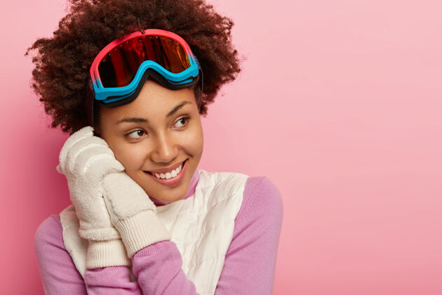 沉思快乐的黑皮肤黑人发型女人 戴着滑雪护目镜 戴着白色柔软的手套 喜欢冬季运动 看起来愉快地离开 孤立在粉红色背景下 感觉好玩和高兴眼镜护目镜一个