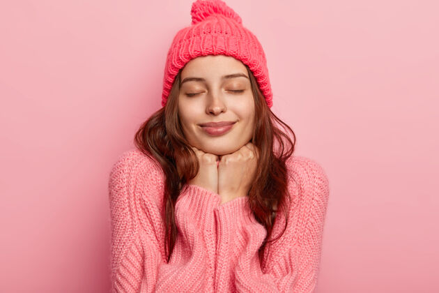 套头衫放松的年轻女模特的画像双手放在下巴下 闭着眼睛 戴着玫瑰色的帽子和套头衫 感觉很满意 在粉色背景下摆姿势寒冷高兴女士