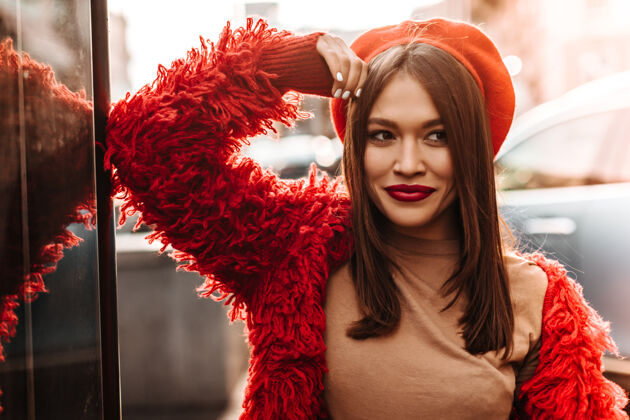 红色夹克一位深色直发 棕色眼睛 头戴红色贝雷帽 身穿环保外套的女士倚在玻璃墙上 在街上摆姿势黑发长发美丽