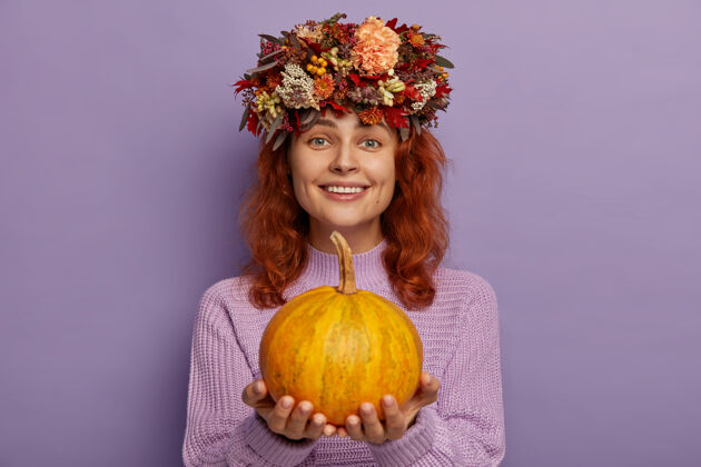 红发漂亮的红发女人戴着秋天的花环 抱着成熟的南瓜 穿着紫色的毛衣装饰室内叶子