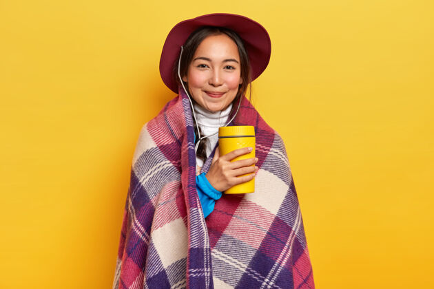 外卖可爱放松的女旅行者喝着热水瓶里的热饮料 站在格子布里 享受徒步旅行 戴着帽子 在黄色背景上摆姿势纸表情持有