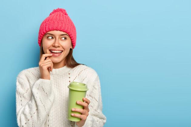 粉色快乐的白种女人的肖像 手指放在嘴唇上 喝外卖咖啡 拿着绿色纸杯 穿着温暖的白色毛衣 专注在一边年轻梦幻女人