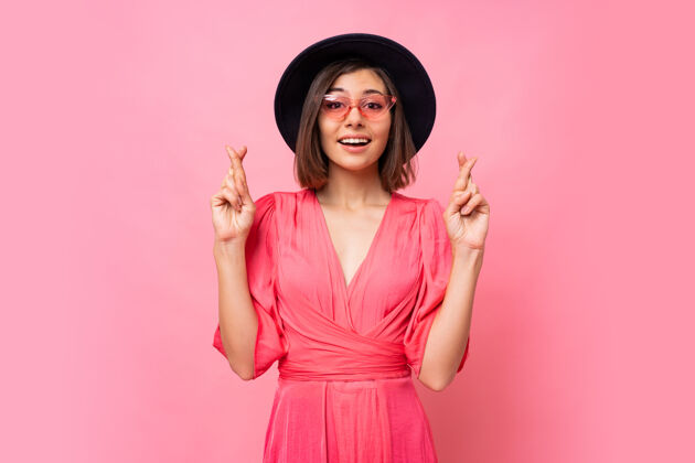 喜悦善良开朗的女人希望尽她最大的努力 让她的手指交叉 拥有最好的 穿着隔离在粉红色墙上的衣服人 肢体语言的概念复制空间室内女孩帽子