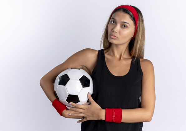 运动装身穿黑色运动服 头戴红色头巾 手持足球 表情严肃的年轻健身女孩站在白色的墙上脸足球女孩