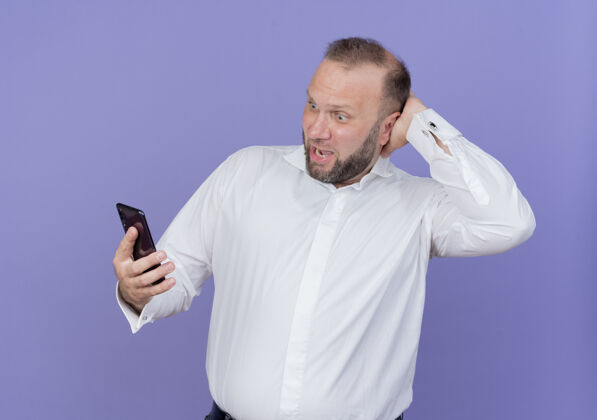 抱着的一个留着胡子的男人穿着白衬衫 手里拿着智能手机 站在蓝色的墙上 看上去既困惑又惊讶惊讶的移动的困惑的