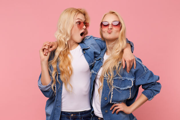站立摄影棚拍摄的年轻快乐迷人的长发金发双胞胎相互取乐和拥抱 而站在粉红色背景下的休闲服女士T恤情绪
