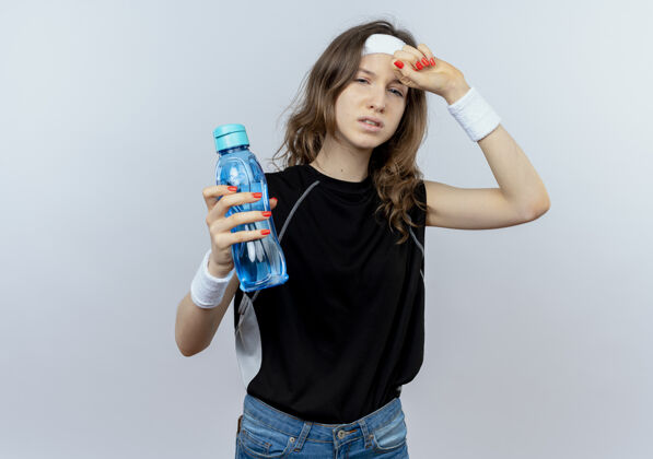 站着穿着黑色运动服的年轻健身女孩 戴着头巾 手里拿着一瓶水 站在白色的墙上 看上去又累又累女孩年轻看着
