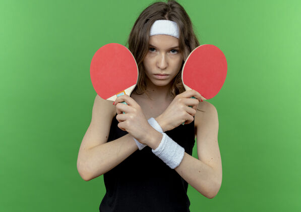 抱身穿黑色运动服的年轻健身女孩戴着头带 手里拿着两个乒乓球拍 不高兴地双手交叉站在绿色的墙上头带乒乓球年轻