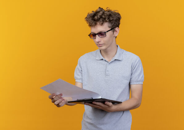 穿一个戴着黑眼镜 穿着灰色马球衫的年轻人拿着一张白纸的剪贴板 严肃地站在橙色的墙上看着它剪贴板严重页