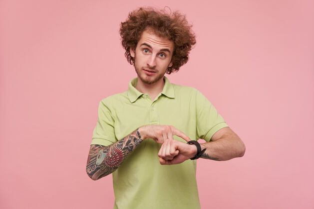 粉色摄影棚照片年轻迷人的卷发黑发男子皱起额头 而显示在手表上与食指 站在粉红色的背景年轻马球墨水