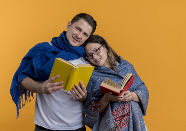 抱着一对年轻夫妇拿着毛毯 抱着书 快乐而积极地微笑着 站在橙色的墙上夫妻女人在一起