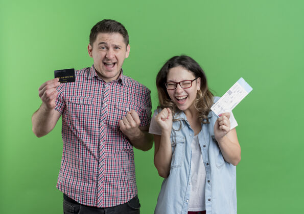 而一对穿着休闲服的年轻男女幸福的男人手里拿着信用卡 而他的女朋友手里拿着机票幸福而兴奋地站在绿色的墙上情侣信用卡兴奋