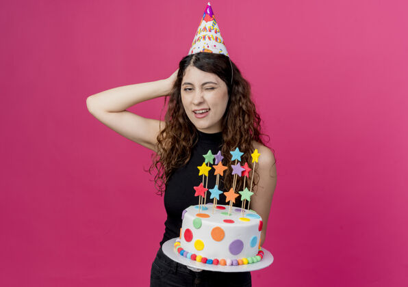 抱着一个年轻漂亮的女人 卷发 戴着节日帽 手里拿着生日蛋糕 头上戴着一只手 站在粉色的墙上 看起来很困惑生日派对的概念年轻困惑蛋糕