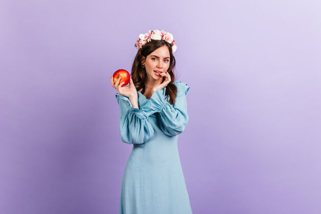 卷发沉思的女孩狡猾地看着 手里拿着开胃的苹果紫丁香墙上戴着花冠的模特肖像服装表情欢呼