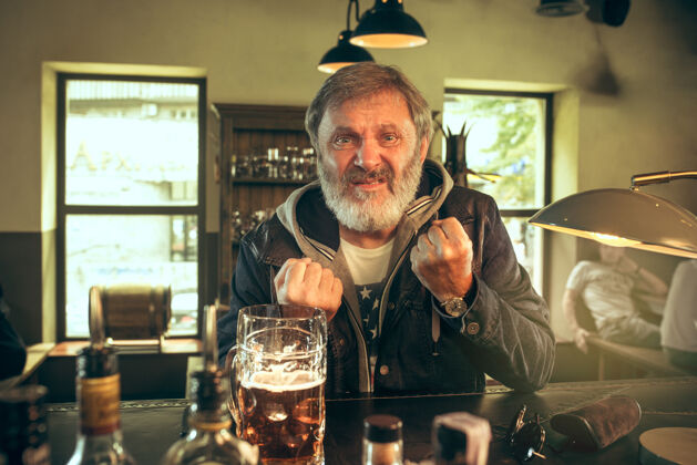 老年人大胡子男人在酒吧喝酒 看电视上的体育节目享受我最喜欢的酒和啤酒男人拿着一大杯啤酒坐在桌边室内酒精坐着
