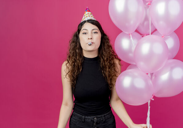 积极一个年轻漂亮的女人 卷发 戴着节日帽 手里拿着一堆气球 快乐而积极地吹着口哨 站在粉色的墙上 庆祝生日气球卷发女人