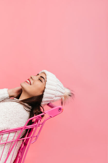 积极可爱的女孩笑着 在粉红色的超市手推车里摆姿势在隔离的墙上 一个身穿白色冬衣的黑发女人的照片年轻女性衣服