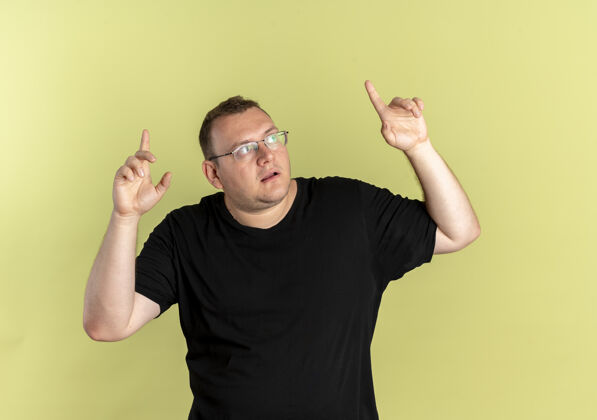 手指一个戴着眼镜 身穿黑色t恤的超重男子抬头困惑地指着 食指站在浅色的墙上超重看站