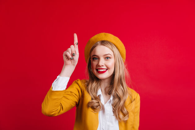 优雅戴贝雷帽的温文尔雅的白人女孩用手指指着迷人的法国女士在红墙上做手势时尚配饰情感