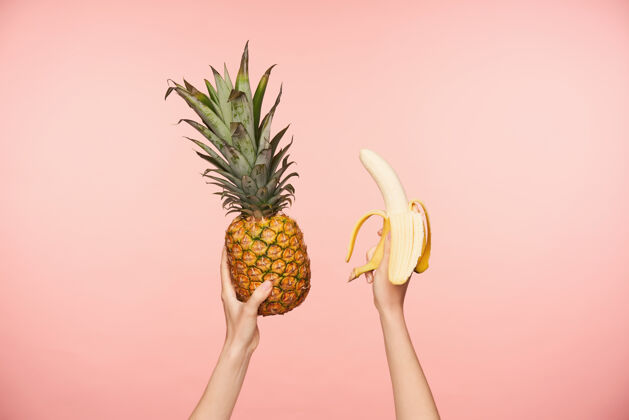 皮肤白皙一张年轻女子手举裸甲的剪影照片 手拿新鲜菠萝和剥皮的香蕉 被隔离在粉色背景上粉红色手室内