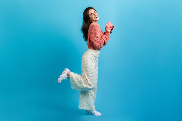针织一个穿着新潮轻装的女孩在蓝色的墙上摆着粉红色的相机一个面带迷人微笑的女士风骚地抬起了她的腿自信有趣女性