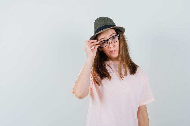 积极年轻女孩透过眼镜看着粉色t恤 戴着帽子 神情犹豫不决 眼前一亮小随意小