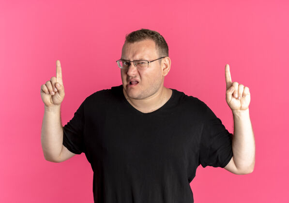 不高兴戴眼镜 身穿黑色t恤的超重男子不高兴地将食指竖立在粉红色的墙上站显示超重