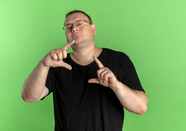佩戴一个戴眼镜的胖男人穿着黑色t恤 手指自信地站在绿色的墙上男人超重长相